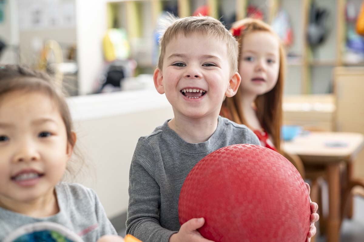 three young children stand next to each other, one of them is holding a red ball