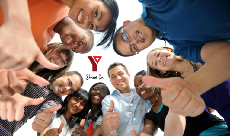 a group of people smile and pose with their thumbs up for a photo above the camera. The center of the photo has the YMCA logo and says "Shine On".